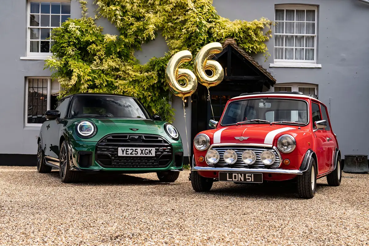 Zwei Generationen MINI vor klassischer britischer Hausfassade: links ein aktueller MINI Cooper S in britischem Racing-Green, rechts ein ikonischer Classic Mini in leuchtendem Rot mit weißen Rallyestreifen und Zusatzscheinwerfern. Zwischen den beiden Modellen goldene Luftballons in Form der Zahl „66“ – ein stilvoller Geburtstagsgruß an die Marke.