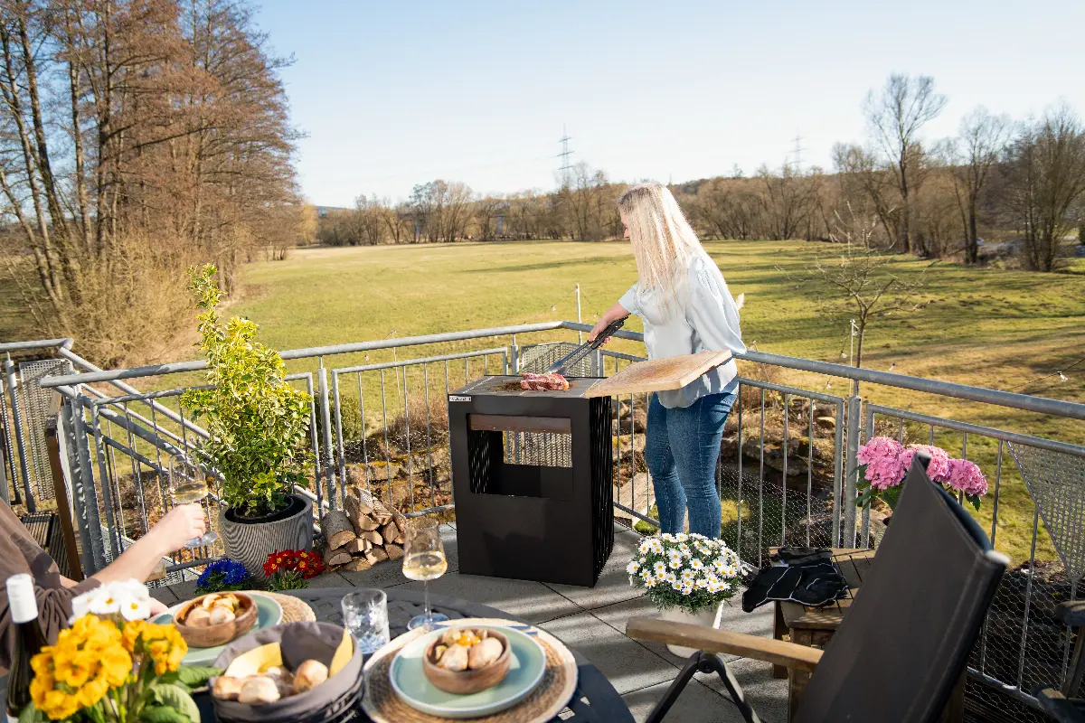 Frau grillt auf dem Outdoorchef FIRE PIT auf einer modernen Terrasse mit Blick ins Grüne, gedeckter Tisch im Vordergrund.